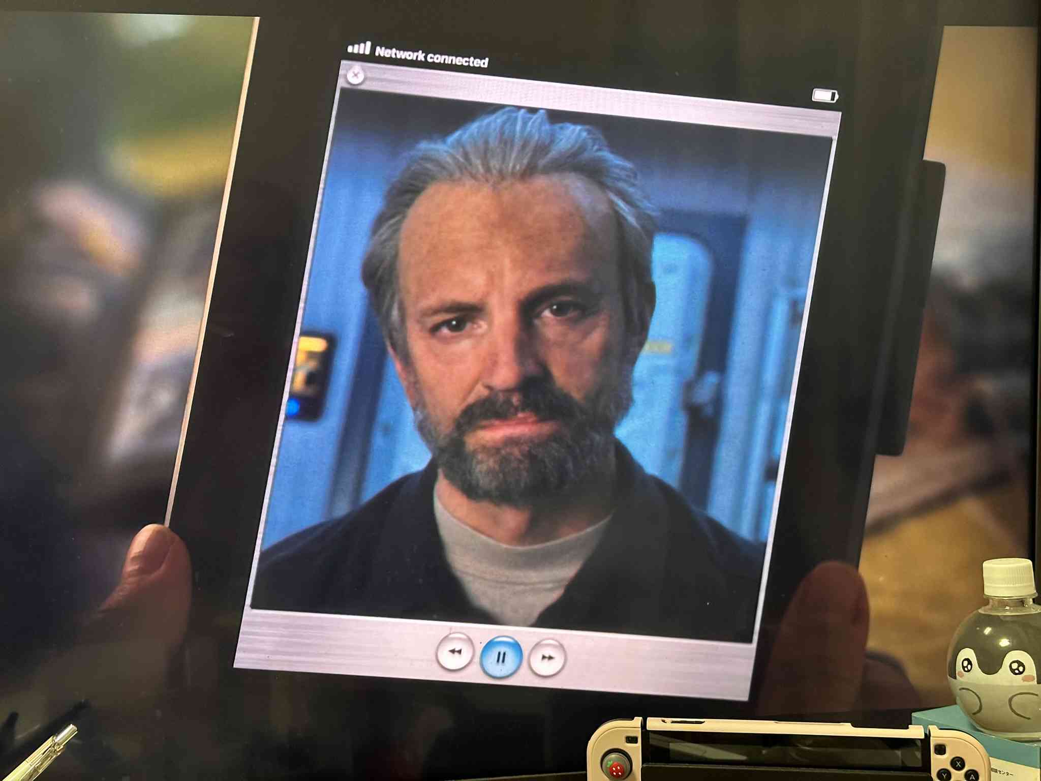 A frame from the TV show For All Mankind showing an iPad-like tablet with a Mac OS X Aqua-style interface, displaying a video feed of an older Edward Baldwin with gray hair and a beard, wearing a dark jacket against a blurred background. Nearby on the desk are a Koupenchan water bottle and a Nintendo Switch