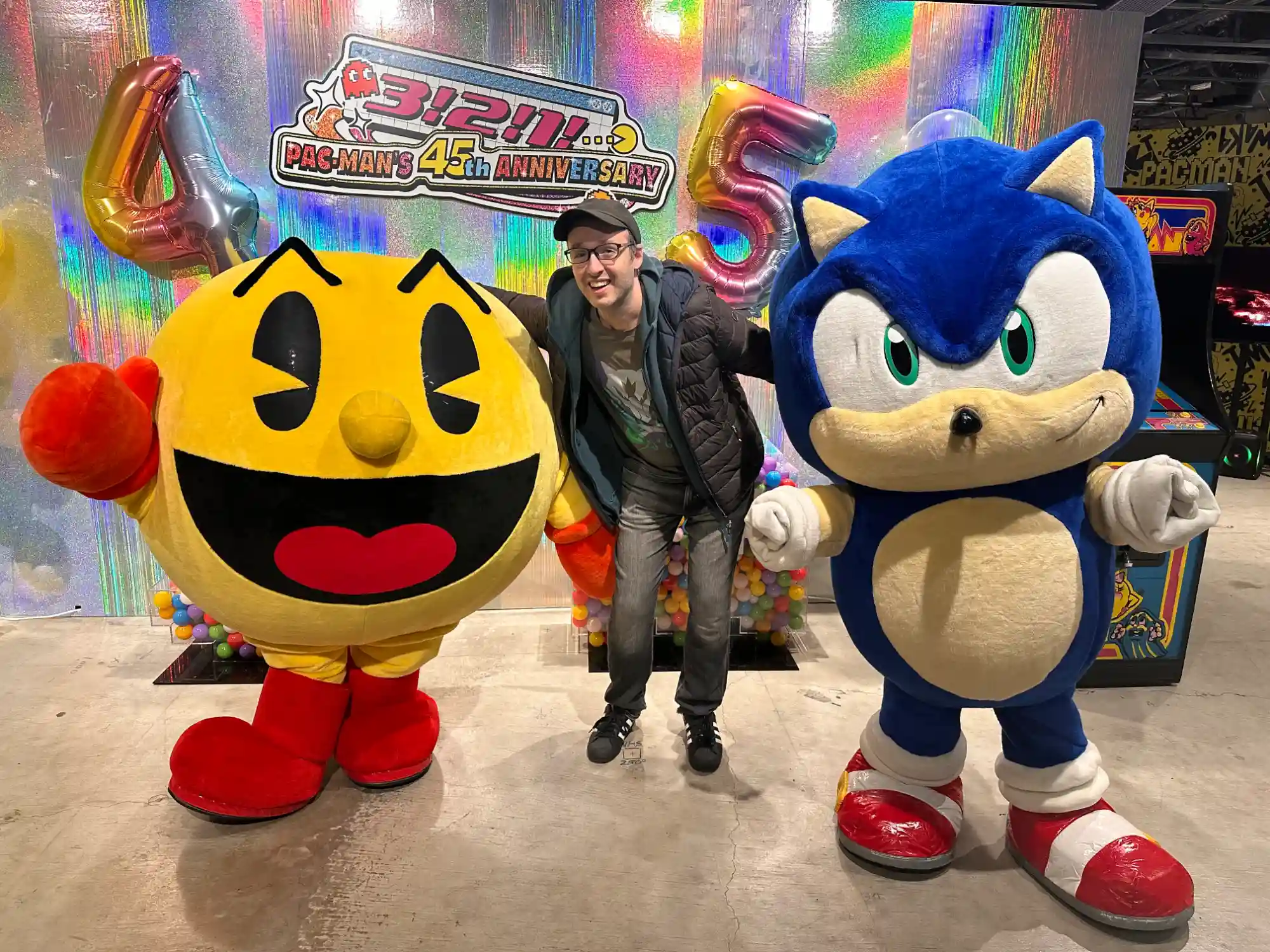 Photo with Pac-Man and Sonic the Hedgehog mascots at the Pac-Man 45th Anniversary event, with a colorful holographic backdrop, balloon numbers, and a Pac-Man arcade cabinet visible on the right