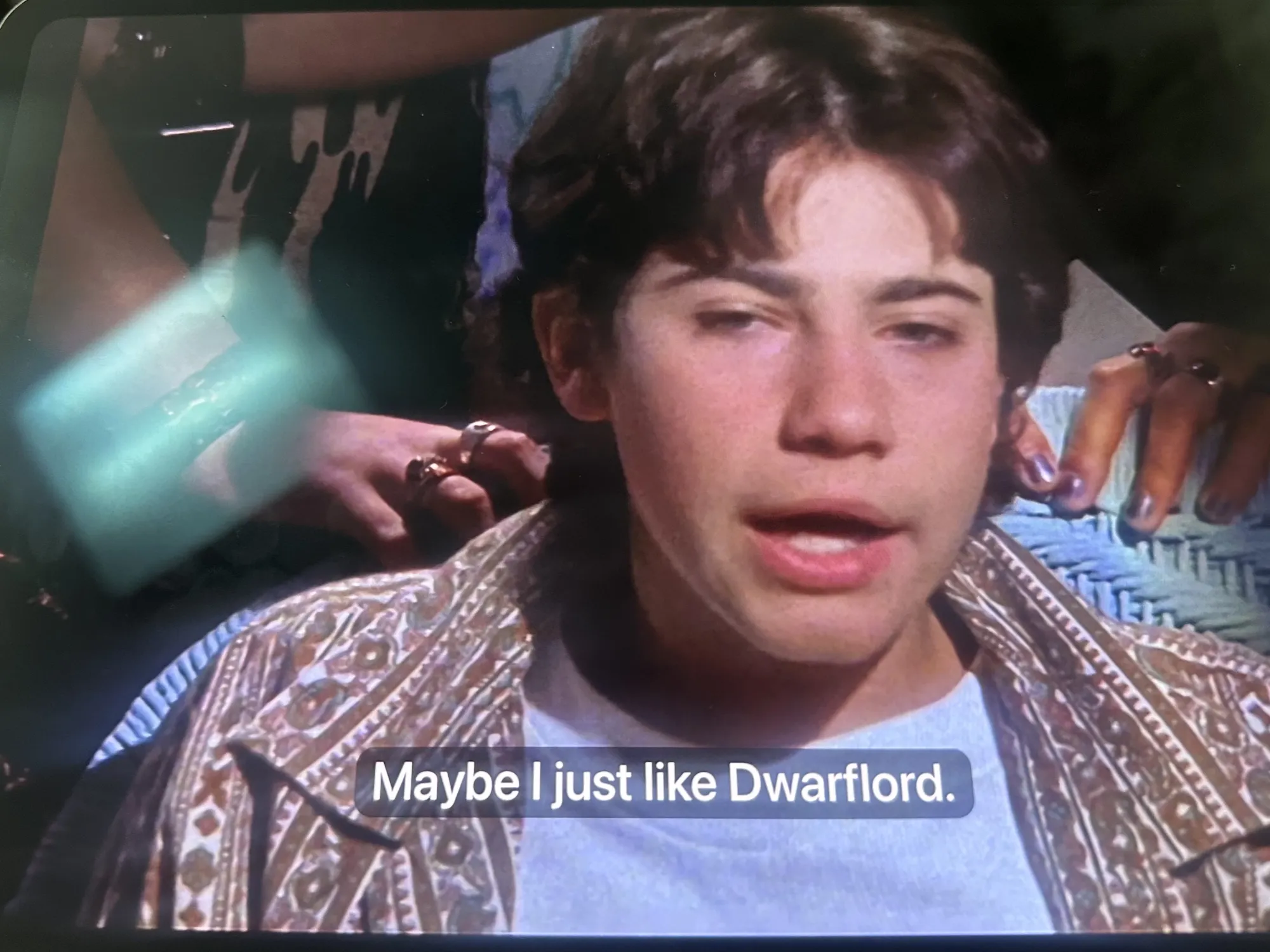 Gordo is wearing a light blue t-shirt underneath an open, intricately patterned brown and white shirt. He has a slightly defensive expression. Overlaid at the bottom of the screen are white subtitles that read, 'Maybe I just like Dwarflord'