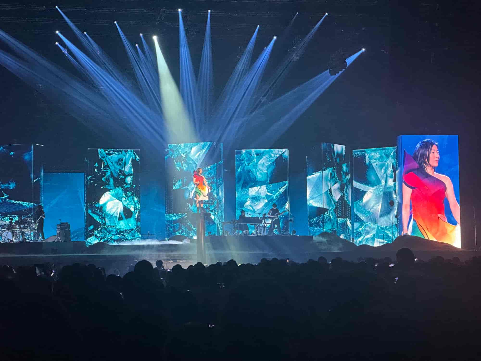 Hikaru Utada performing on a vibrant stage surrounded by tall LED screens displaying dynamic blue visuals and a close-up of them in a colorful outfit. Bright beams of light illuminate the scene, creating an immersive atmosphere as the audience watches in awe